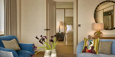 Living area of a suite at The River Lee hotel with a sofa, armchair, fresh flowers, and a king bed visible through an open door.