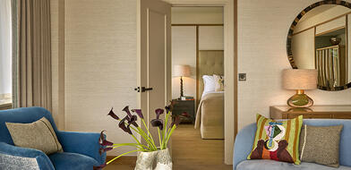 Living area of a suite at The River Lee hotel with a sofa, armchair, fresh flowers, and a king bed visible through an open door.
