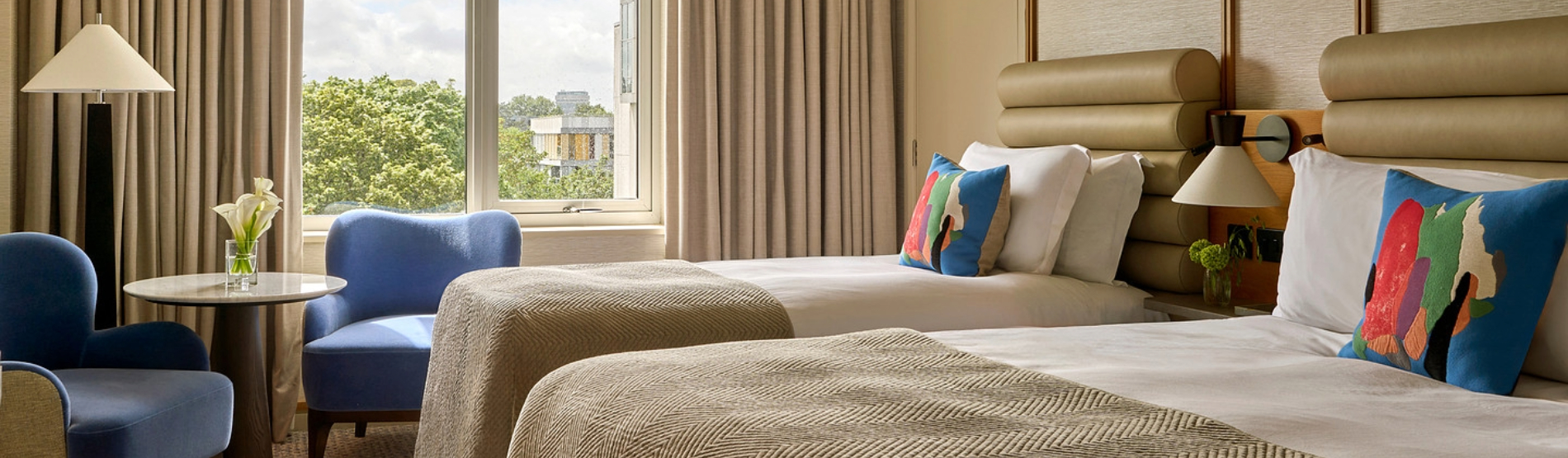 Guest room at The River Lee with a deluxe double bed, a single bed, crisp white linens, and two blue armchairs with a small side table.