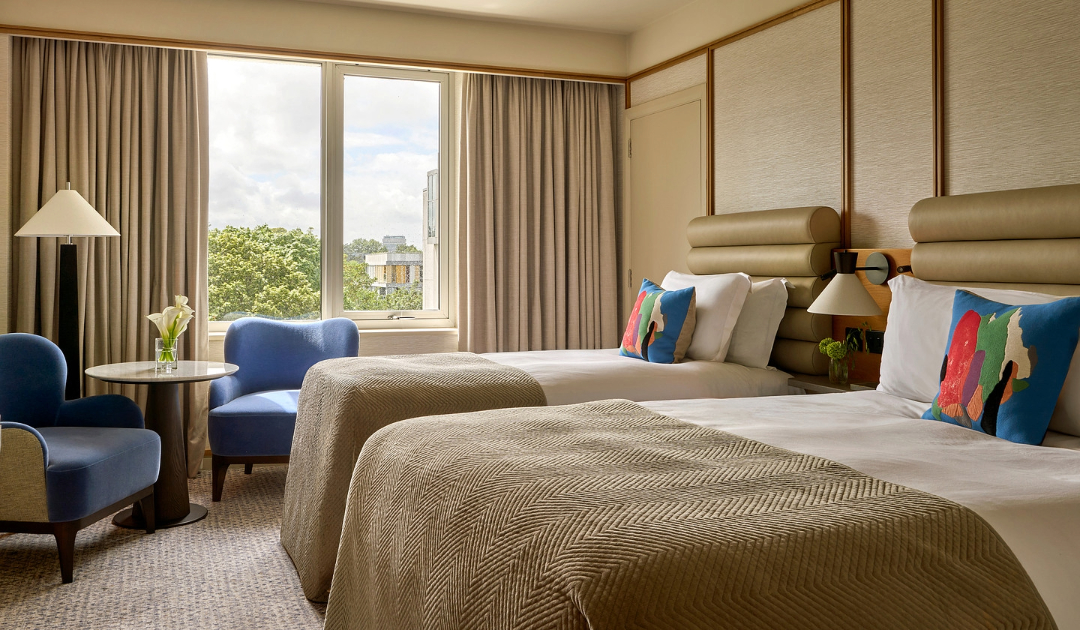 Guest room at The River Lee with a deluxe double bed, a single bed, crisp white linens, and two blue armchairs with a small side table