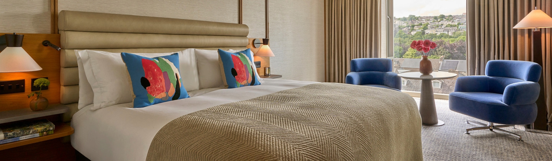 A vibrant, guest room at The River Lee hotel with river view, featuring a large deluxe bed with crisp white linens flanked by two modern blue armchairs and a small side table.