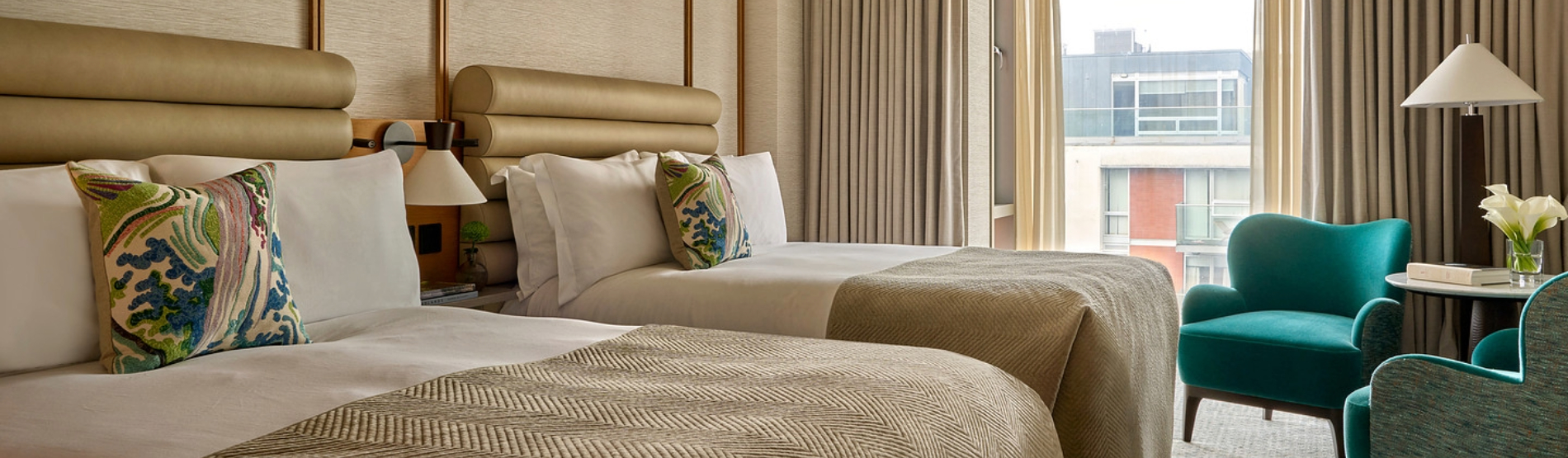 A spacious guest room at The River Lee showcasing two deluxe double beds with clean white linens, complete with two stylish blue armchairs and a small side table for group or family stays.
