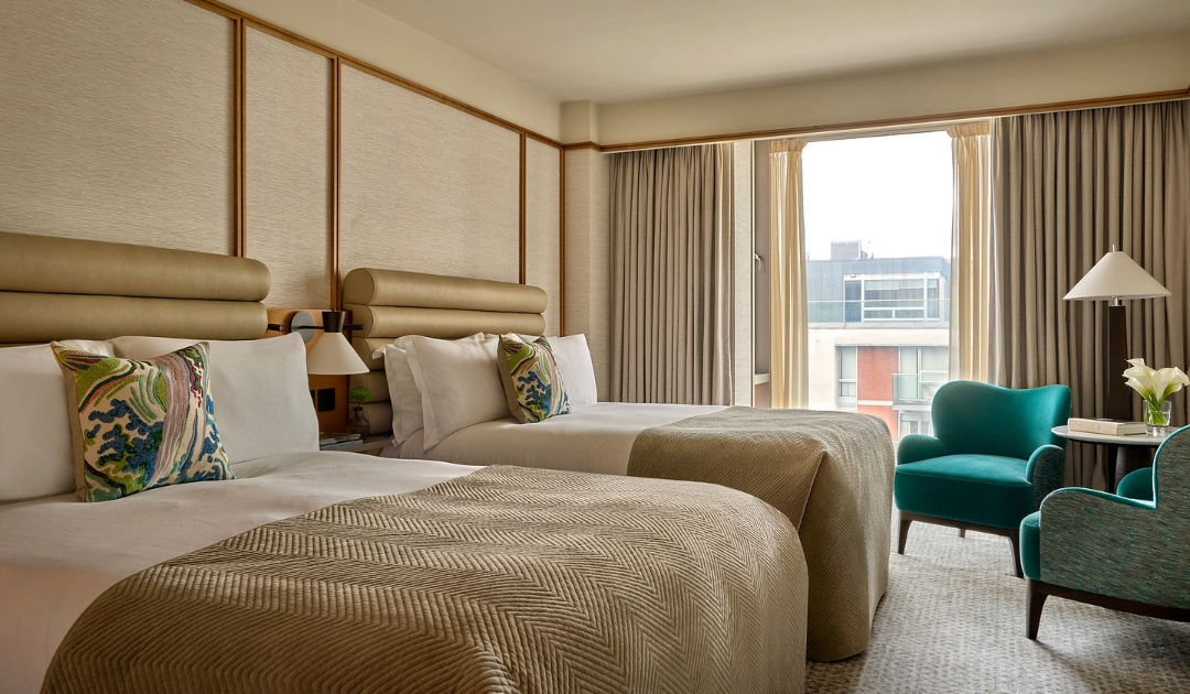 A spacious guest room at The River Lee showcasing two deluxe double beds with clean white linens, complete with two stylish blue armchairs and a small side table for group or family stays.