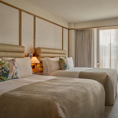 Deluxe Room with two double beds at The River Lee Hotel, featuring large windows, modern decor, and two signature blue armchairs, ideal for a family or group stay.