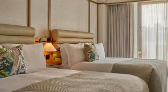 Deluxe Room with two double beds at The River Lee Hotel, featuring large windows, modern decor, and two signature blue armchairs, ideal for a family or group stay.