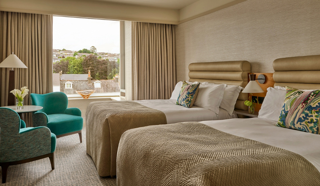 A river-view Classic Room at The River Lee Hotel with two double beds, a lampshade, and two turquoise armchairs around a small coffee table