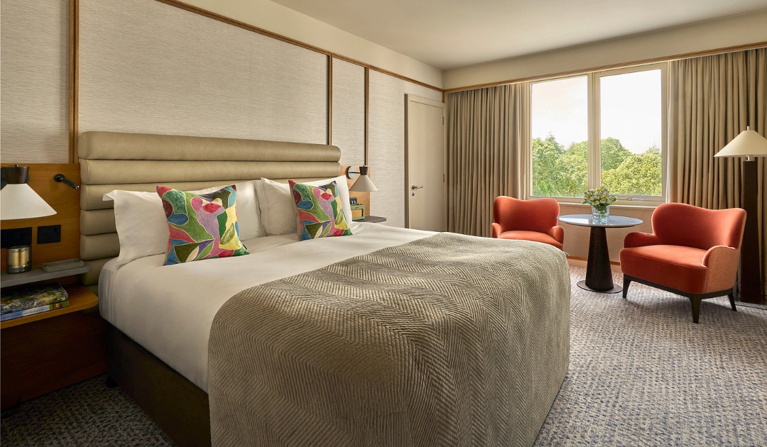 A spacious, modern hotel room with a king-sized bed, a large window overlooking greenery, and a small table and two armchairs
