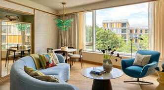 llustrative view of the Luxury Suite seating area at The River Lee, featuring a large sofa bed, armchair, and a 4-person dining table in a bright, sunlit room