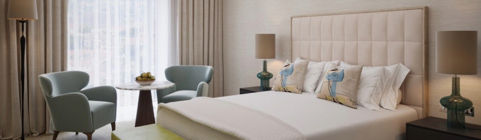 Illustrative view of a light-filled luxury bedroom featuring a king bed and a cozy seating area with two chairs.