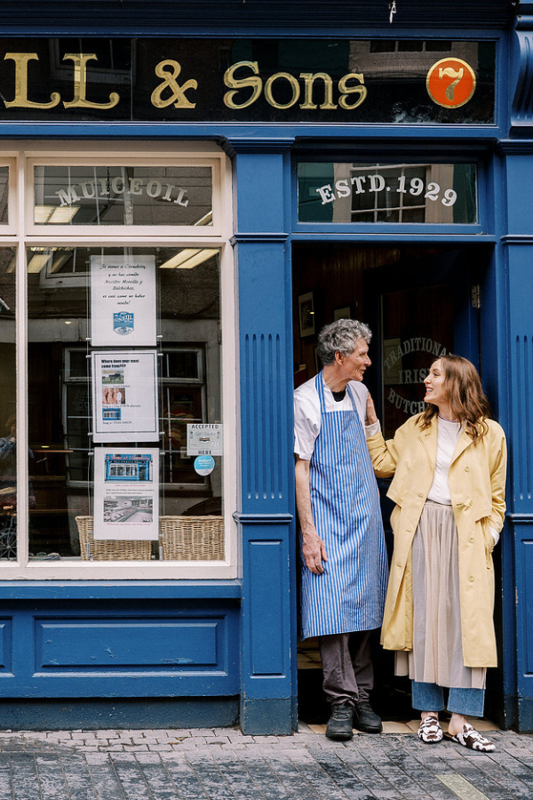 Louise and her father sit together in the doorway of O’Neill’s butcher shop, sharing a warm smile in a relaxed family moment.