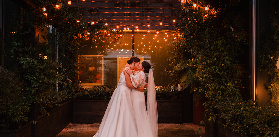 Sinead & Laura embrace and kiss surrounded by terrace greenery 