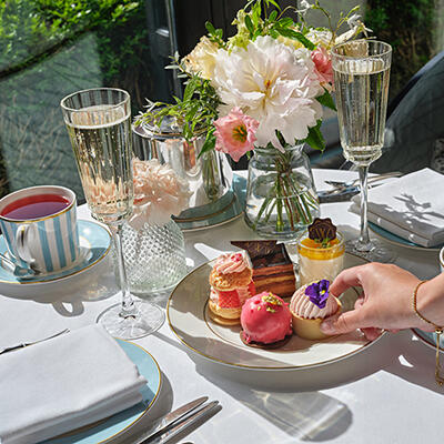 An elegant afternoon tea setting at The Gallery, featuring champagne flutes, a teacup, and a plate of colorful pastries, with a hand selecting a dessert.