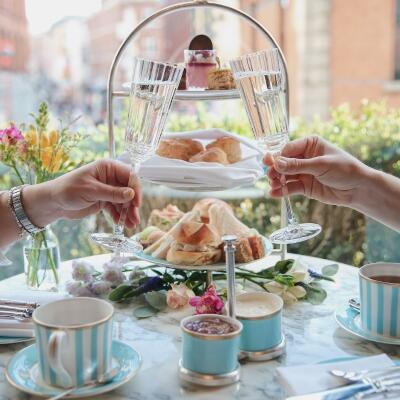 A celebratory toast with two champagne flutes clinking in front of a window at The Westbury, with a tiered afternoon tea stand blurred in the background