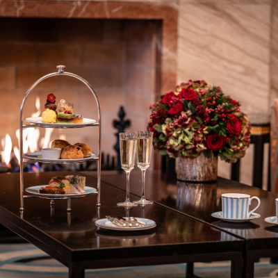 Elegant afternoon tea setting at The Galery, with two champagne flute and a tiered stand filled with various pastries and sandwiches set beside a warmly lit fireplace and festive Christmas flowers.