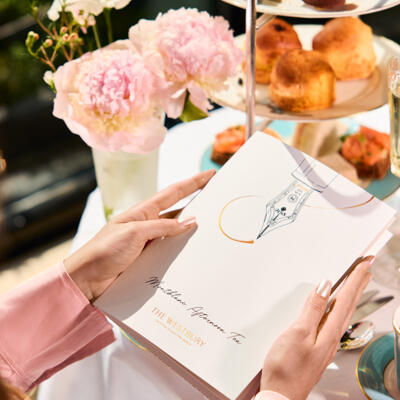 An elegant afternoon tea setting in The Gallery, featuring a beautifully dressed table with a selection of afternoon tea pastries, fresh flowers, and a guest browsing the menu, creating a refined and inviting atmosphere.