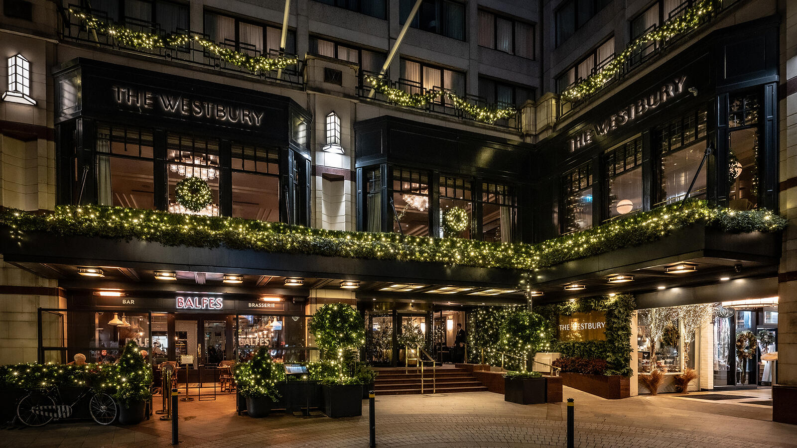 The exterior of The Westbury Hotel is brightly lit at night, showcasing Christmas decorations, including string lights and wreaths, creating a festive holiday scene.