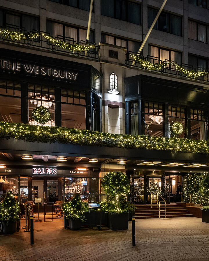 The exterior of The Westbury Hotel is brightly lit at night, showcasing Christmas decorations, including string lights and wreaths, creating a festive holiday scene.