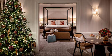 Luxurious suite at The Westbury hotel, featuring a four-poster bed and a decorated Christmas tree. A table is set with festive flowers and two glasses of Champagne.