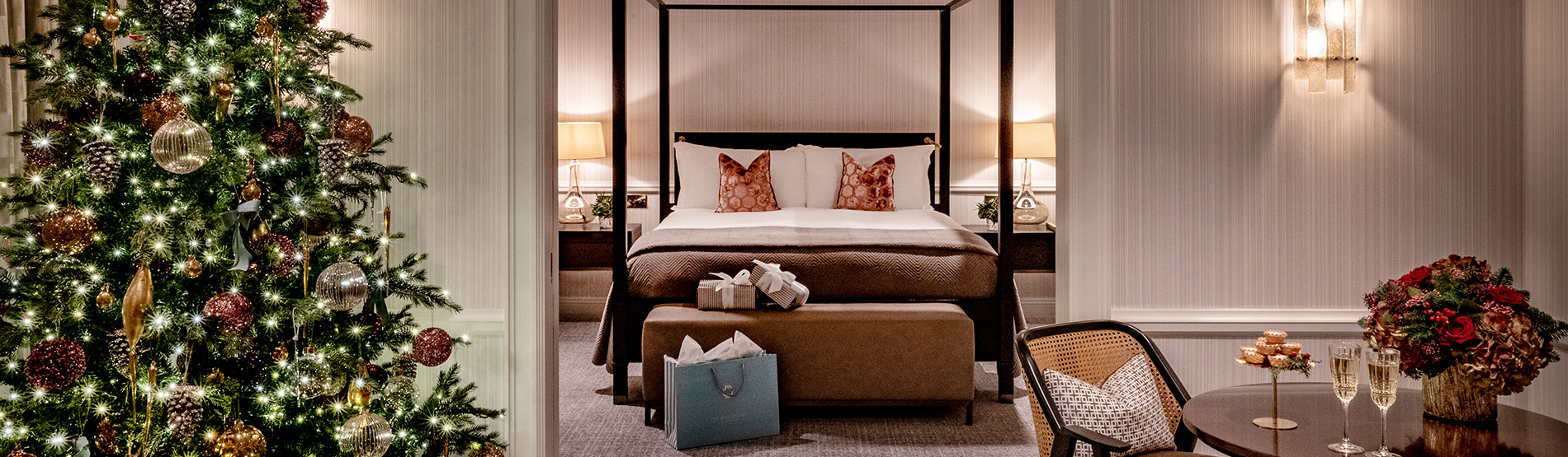 Luxurious suite at The Westbury hotel, featuring a four-poster bed and a decorated Christmas tree. A table is set with festive flowers and two glasses of Champagne.