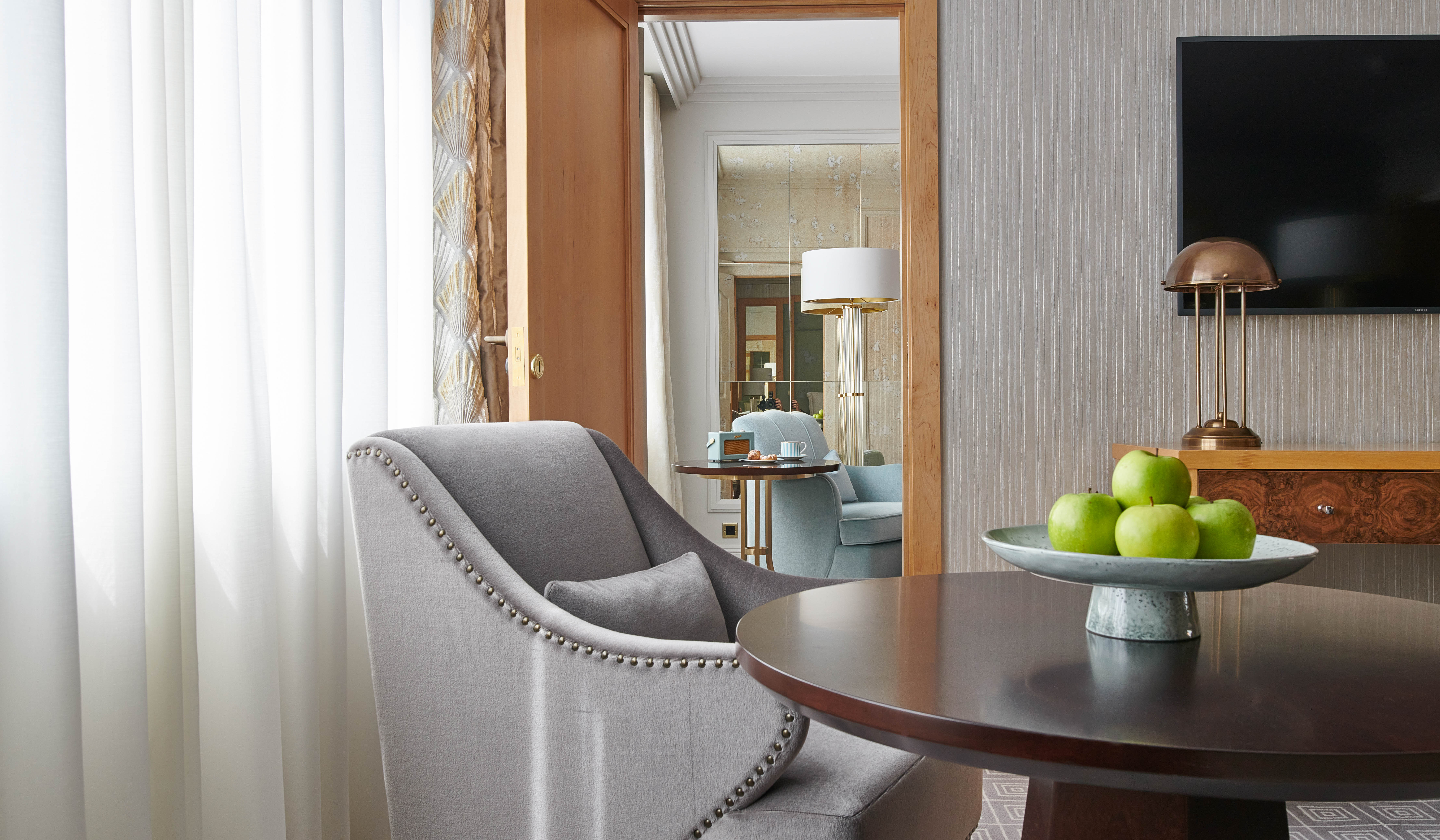 A bright, interconnecting luxury suite at The Westbury, showing an open doorway. The foreground features a stylish lounge table with fresh green apples, leading into a second room with a prepared coffee set, emphasizing a spacious family-style layout
