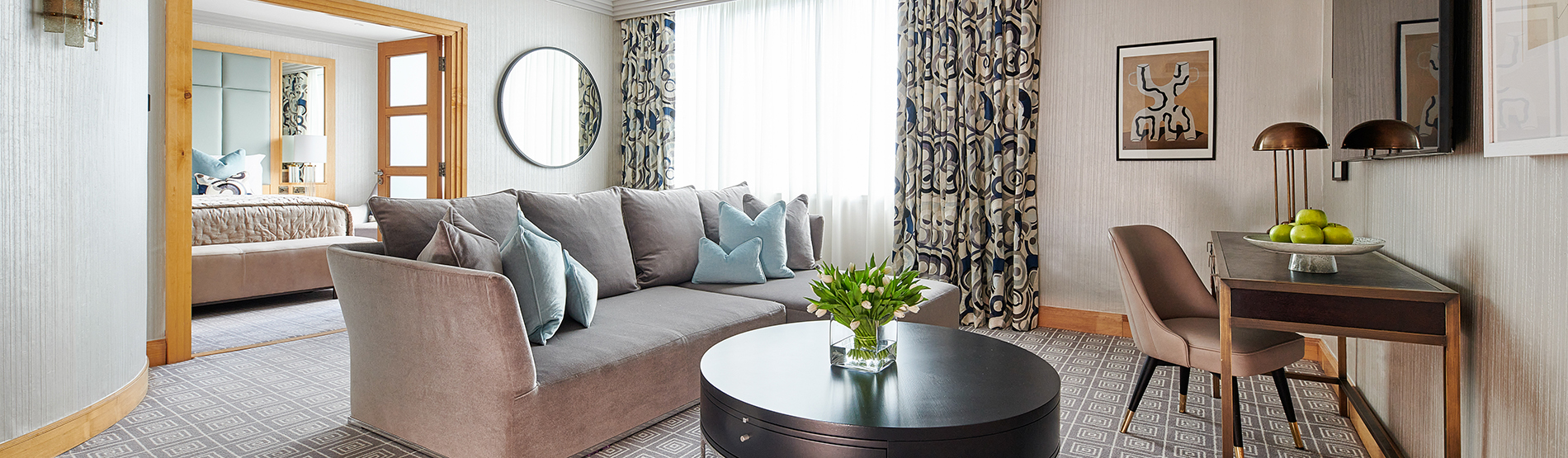 The spacious living area of a luxury three-bedroom suite at The Westbury. The room features a plush sofa and a coffee table adorned with fresh flowers, offering a glimpse into the master bedroom through an open doorway.