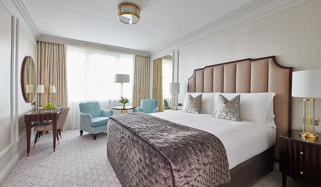 A master bedroom in a three-bedroom suite at The Westbury, featuring a king-size bed and two elegant light blue armchairs positioned by a bright window.