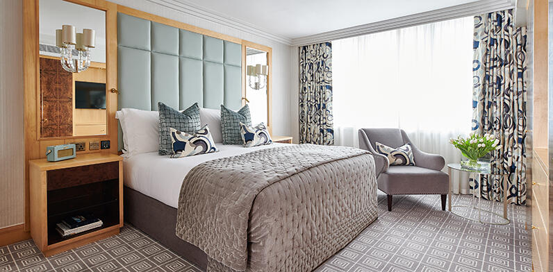 Luxury suite master bedroom at The Westbury, showcasing contemporary grey and light blue interiors, a king-size bed, a stylish armchair, and fresh floral accents