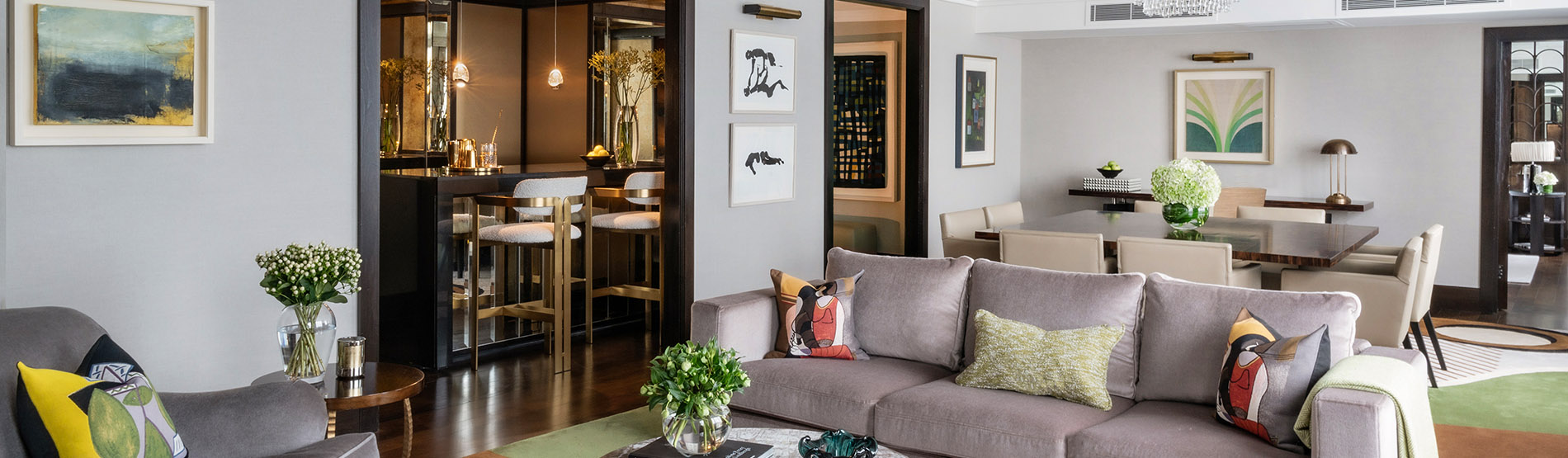A spacious living room with a grey sofa, a coffee table, and a dining table with chairs. There is a small bar area with stools and a wine rack. The walls are decorated with artwork and there are plants and flowers throughout the room.