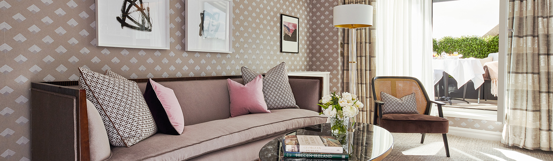 The living room of the Terrace Suite at The Westbury Hotel, featuring elegant furnishings, dynamic wallpaper, and statement artwork. The space is spacious and tastefully designed, with a comfortable sitting area that invites relaxation. Large windows provide a view of the fully furnished private terrace, creating a seamless blend of indoor and outdoor luxury.